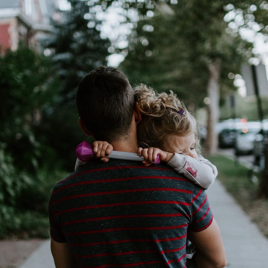 tired little girl rests her head on her father's shoulder as he carries her down the city sidewalk