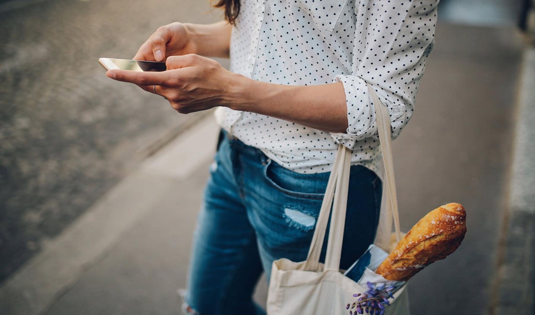 woman walks down the street and texts with a bag of groceries on her arm