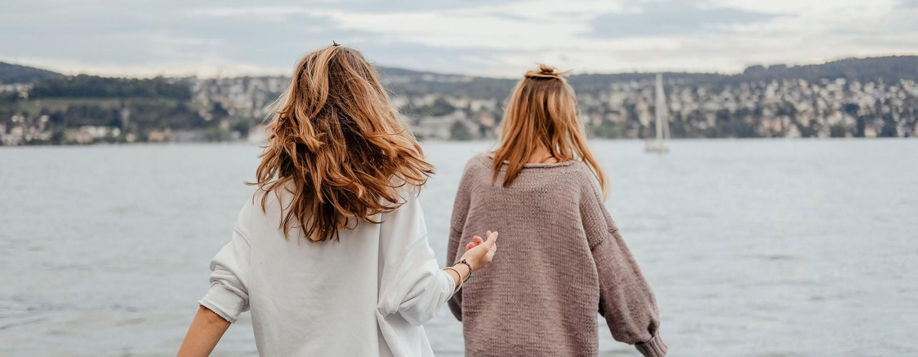 two women walk to the edge of a dock, in a body of water, looking toward the city on the other shore